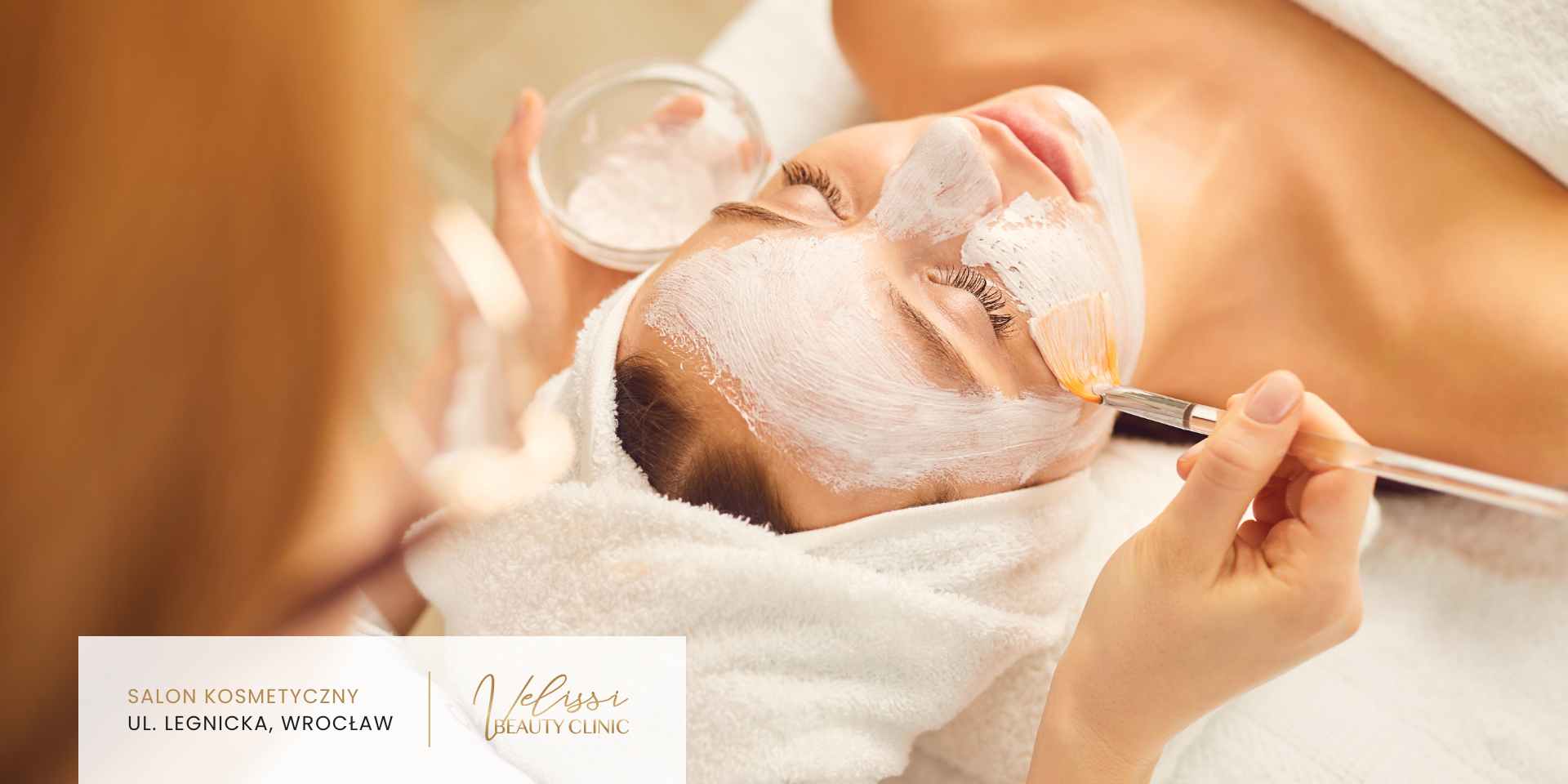 peeling-chemiczny-efekty-wroclaw Klientka w białym ręczniku na głowie leży na leżance, podczas gdy kosmetolog pędzlem nakłada na jej twarz peeling chemiczny.