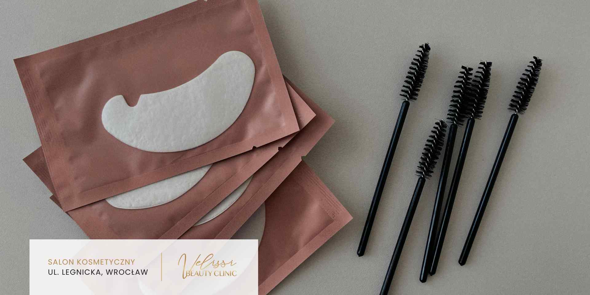 Na jasnym tle leżą cztery czarne szczoteczki do rzęs oraz trzy różowe saszetki z białymi płatkami pod oczy.